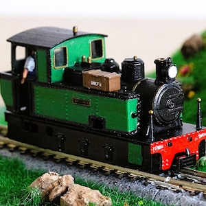 May include: A green and black toy train engine with a small figure of a person in the cab. The engine has a brown box on top and the text "SN.CF.R" on the side. The train is on a model railroad track.