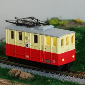 May include: A model train car with a red and cream-colored body, a gray roof, and a black undercarriage. The train car has illuminated windows and is positioned on a model train track. The number 20 is visible on the side.