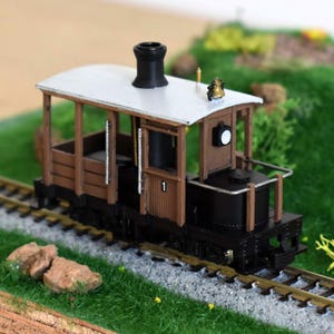 Könnte beinhalten: Ein Miniatur-Modellzug mit braunem und schwarzem Gehäuse, weißem Dach und schwarzem Schornstein. Der Modellzug befindet sich auf einer Miniaturstrecke mit grünem Gras und kleinen Felsen. Die Nummer "1" ist auf der Seite sichtbar.
