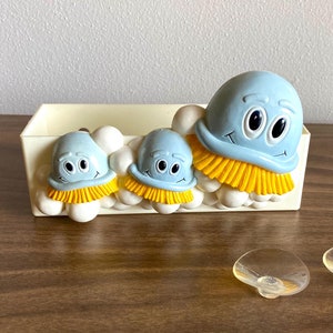 May include: A whimsical, light blue jellyfish-themed decorative box with three smiling jellyfish figures. The jellyfish have yellow fringe and are set against a white background. Two clear, mushroom-shaped objects are in front of the box.