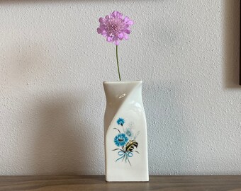 May include: A white ceramic vase with a blue floral design and a single pink flower. The vase has a unique, twisted shape and is placed on a wooden surface. Artwork hangs in the background.