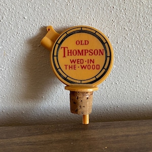 May include: A yellow plastic bottle stopper with a cork top. The stopper is shaped like a barrel and has the text "Old Thompson Wed-In The Wood" printed on it.