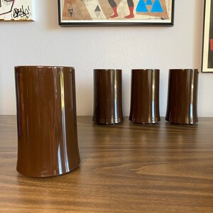 May include: Four brown glass tumblers on a wooden surface. The tumblers are all the same size and shape, and they have a smooth, rounded bottom.