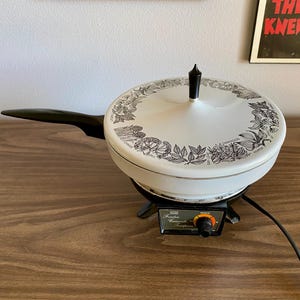 May include: Vintage electric skillet with a white lid featuring a black botanical design. The skillet has a black handle and sits on a black base with a temperature control dial. A power cord extends from the base.