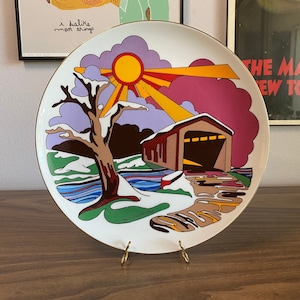 May include: A decorative plate with a colorful winter scene. The design features a covered bridge, a tree, and a sunburst. The plate has a gold rim and is displayed on a gold stand.