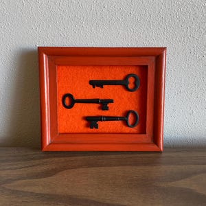 May include: Three antique keys displayed in a red wooden frame with an orange felt background.