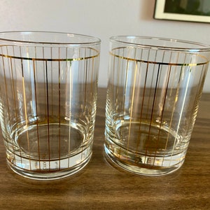 May include: Two clear glass tumblers with gold stripes. The glasses are empty and sitting on a wooden surface.
