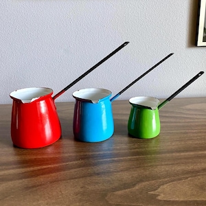 May include: Three colorful enamel coffee pots in red, blue, and green, each with a white interior and a long, black handle. The pots are arranged in a row on a wooden surface, showcasing their vibrant colors and classic design.