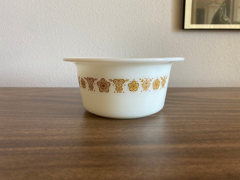 May include: White ceramic bowl with a gold floral pattern around the rim. The bowl is on a brown wooden surface.