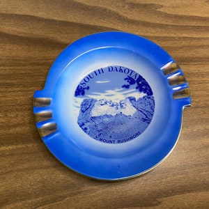 May include: Blue and white ceramic ashtray with a gold rim. It features a picture of Mount Rushmore with the text "South Dakota Mount Rushmore".