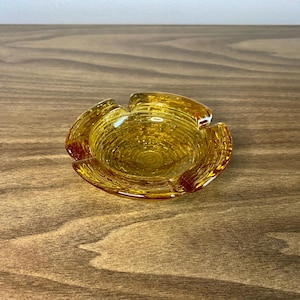 May include: A small, amber-colored glass ashtray with a textured surface and a four-leaf clover design.