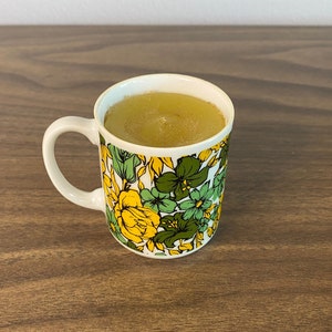 May include: A white mug with a floral pattern in shades of yellow, green, and brown. The mug is filled with a light yellow liquid.