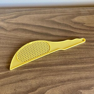 May include: A yellow plastic kitchen utensil with a perforated, curved head and a handle. The utensil is designed for straining or draining food items. The handle has a small hole for hanging.