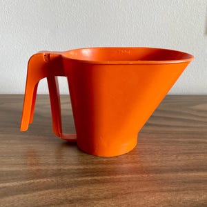 May include: Bright orange plastic measuring cup with a handle. The cup has a wide opening and a spout for pouring. The side of the cup has a measurement marking of 750 ml.