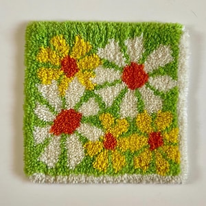 May include: Square, handmade rug with a floral design. The rug features a green background with yellow, white, and orange daisy-like flowers. The flowers are arranged in a repeating pattern. The rug has a white border.