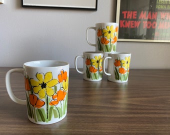 May include: Four white ceramic mugs with a floral design featuring yellow, orange, and green flowers. The mugs have a handle on the right side.
