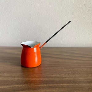 以下が含まれることがあります： 鮮やかなオレンジ色のエナメル製コーヒーポットで、内側は白く、長く細い黒いハンドルが付いています。ポットには小さな注ぎ口があり、滑らかで光沢のある仕上がりです。ハンドルはポットの側面に取り付けられ、斜め上に向かって伸びています。