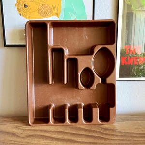 May include: A brown plastic utensil organizer with various compartments for silverware. The organizer has slots for forks, knives, and spoons, along with other sections for serving utensils. The rectangular tray is designed to fit inside a drawer.