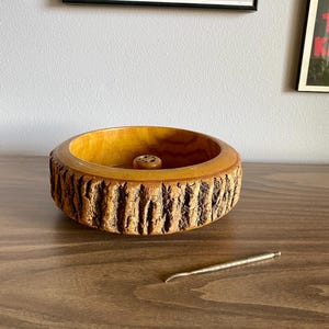 May include: A wooden nut bowl with a textured bark-like exterior and a small, round nutcracker. A silver nut pick rests on the wooden surface. The bowl is light brown, and the pick is silver.