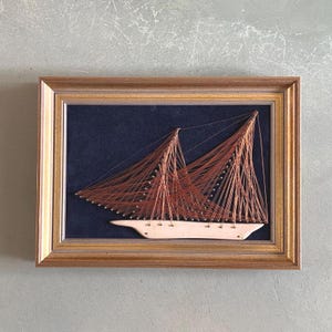 May include: A framed string art piece depicting a sailboat with a brown wooden frame and a navy blue background.
