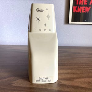 May include: Cream-colored Oster ice crusher with the brand name and starburst designs. The word "ICER" is printed below the logo. A label at the bottom reads "CAUTION KEEP FINGERS OUT".