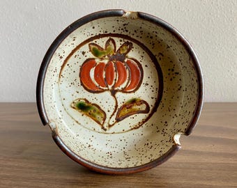 May include: A ceramic ashtray with a floral design. The ashtray is round with a speckled cream-colored interior and a dark brown rim. A red and orange flower with green leaves is painted in the center.