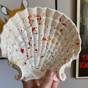 May include: A white ceramic seashell dish with red, yellow, and blue paint splatters. The dish is shaped like a scallop shell and has a slightly raised rim.
