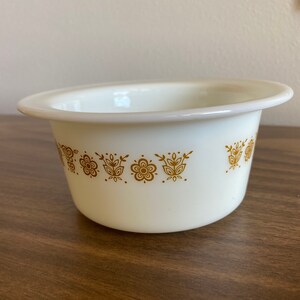 May include: White ceramic bowl with a gold floral pattern around the rim. The bowl is on a wooden surface.