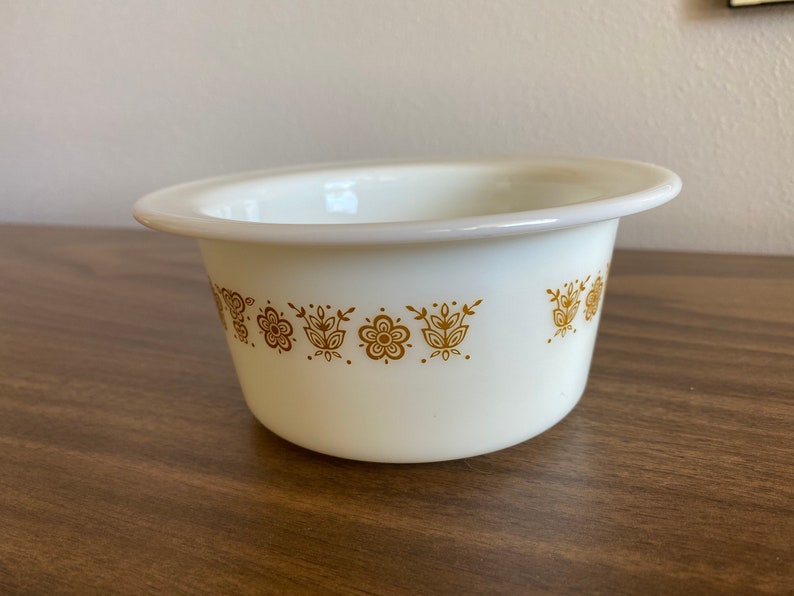 May include: White ceramic bowl with a gold floral pattern around the rim. The bowl is on a wooden surface.