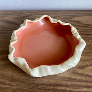 May include: A decorative ceramic bowl with a ruffled edge. The interior is a soft coral color, and the rim is a creamy off-white. The bowl is sitting on a wooden surface.