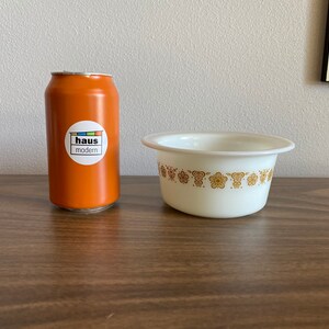 May include: A white ceramic bowl with a gold floral pattern around the rim. The bowl is sitting on a brown wooden surface next to an orange can with a "haus modern" sticker.