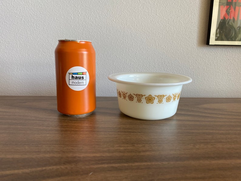 May include: A white ceramic bowl with a gold floral pattern around the rim. The bowl is sitting on a brown wooden surface next to an orange can with a "haus modern" sticker.