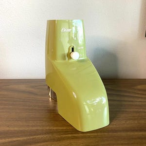 May include: A green Oster electric ice crusher with a white knob and a clear plastic container. The crusher is on a wooden surface.