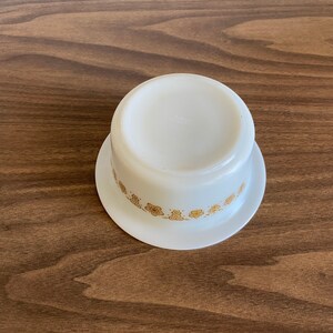 May include: White ceramic bowl with a gold floral pattern around the rim. The bowl is sitting on a wooden surface.