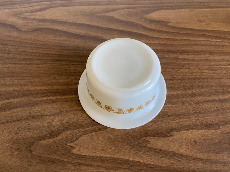 May include: White ceramic bowl with a gold floral pattern around the rim. The bowl is sitting on a wooden surface.