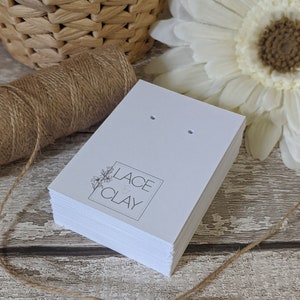Puede incluir: Una pila de tarjetas blancas para pendientes con dos agujeros para exhibir pendientes. Las tarjetas tienen un logotipo en blanco y negro que dice "Lace & Clay" con un peque&ntilde;o dise&ntilde;o floral.
