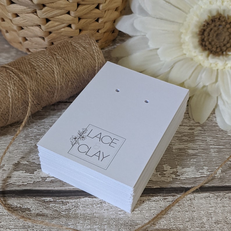 Puede incluir: Una pila de tarjetas blancas para pendientes con dos agujeros para exhibir pendientes. Las tarjetas tienen un logotipo en blanco y negro que dice "Lace & Clay" con un peque&ntilde;o dise&ntilde;o floral.