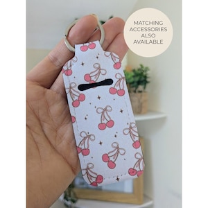 May include: A white keychain holder with a pattern of pink cherries and brown bows. The holder has a silver key ring and a black opening. Text on the image says "MATCHING ACCESSORIES ALSO AVAILABLE."