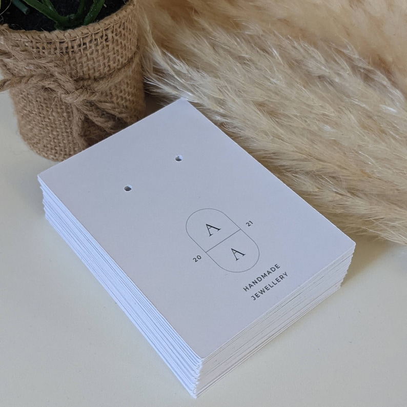 Puede incluir: Una pila de tarjetas blancas para pendientes con dos agujeros para pendientes. Las tarjetas tienen un logotipo con las letras "A" en un c&iacute;rculo y el texto "HANDMADE JEWELLERY".