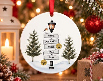 May include: A round white ceramic Christmas ornament hanging from a red ribbon, featuring a black street lamp post with five signs labelled Paul, Lorraine, Mike, Izzy, and Sam, two pine trees, a small bird, and two gold circular pet tags.