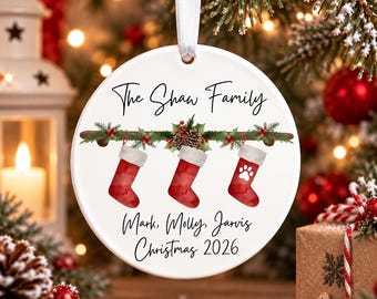 May include: A round white ceramic ornament hanging from a white ribbon. It features a watercolor illustration of three red stockings hanging from a pine branch with pinecones and berries. Text reads: The Shaw Family, Mark, Molly, Jarvis, Christmas 2026.