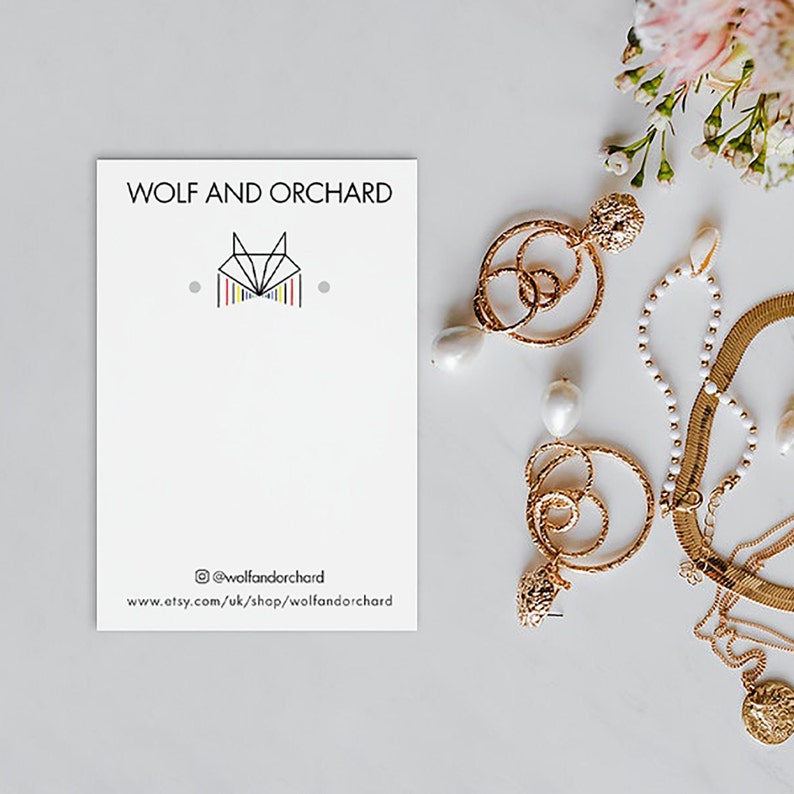 Puede incluir: Una tarjeta blanca con el texto "WOLF AND ORCHARD" y un logotipo de zorro estilizado. La tarjeta est&aacute; rodeada de joyas de oro, incluyendo aretes de aro con acentos de perlas, un collar de cadena de oro y una pulsera de oro.