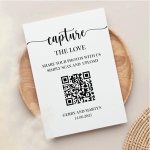 May include: A white sign with black text: "capture THE LOVE SHARE YOUR PHOTOS WITH US SIMPLY SCAN AND UPLOAD." A QR code is below the text, and the names "GERRY AND MARTYN" and the date "14.06.2025" are at the bottom. The sign is on a wooden plate.