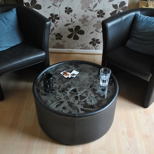 Puede incluir: Dos sillones de cuero negro con cojines azules se enfrentan a través de una mesa de café redonda con tapa de vidrio. La tapa de la mesa presenta un diseño de engranajes y piñones.