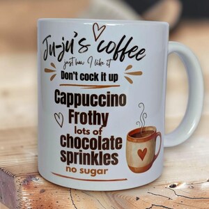 May include: A white ceramic mug featuring the black and brown text Ju-ju's coffee just how I like it Don't cock it up Cappuccino Frothy lots of chocolate sprinkles no sugar, with a small illustration of a mug with a heart and steam.