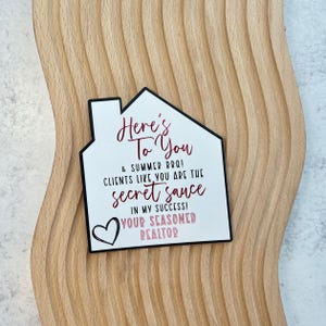 May include: A white house-shaped sign with black trim and the text "Here's To You & Summer BBQ! Clients like you are the secret sauce in my success! Your Seasoned Realtor" in red script. A heart is in the lower left corner.