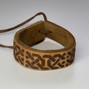 Puede incluir: Un brazalete de cuero marrón con un diseño de nudo celta. El brazalete está atado con un cordón marrón.