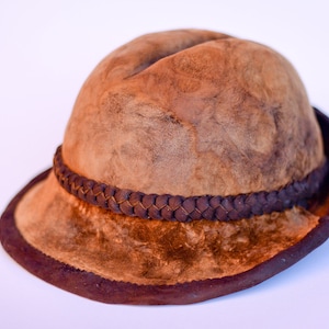 Puede incluir: Un sombrero de cuero marrón con una banda de cuero trenzada. El sombrero tiene un ala ancha y una corona redondeada.