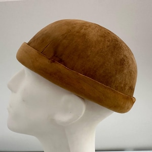 Puede incluir: Un sombrero marrón redondeado con un borde enrollado. El sombrero está hecho de un material texturizado y se asienta sobre una cabeza de maniquí blanca. El color del sombrero es un tono cálido y terroso, lo que sugiere un estilo vintage o rústico.