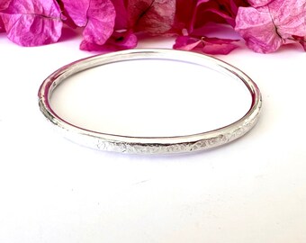 Puede incluir: Una pulsera rígida de plata con una superficie interior lisa y un acabado exterior martillado y texturizado. La banda de metal es circular y reflectante, mostrando un brillo metálico que capta la luz en su superficie exterior irregular y trabajada.